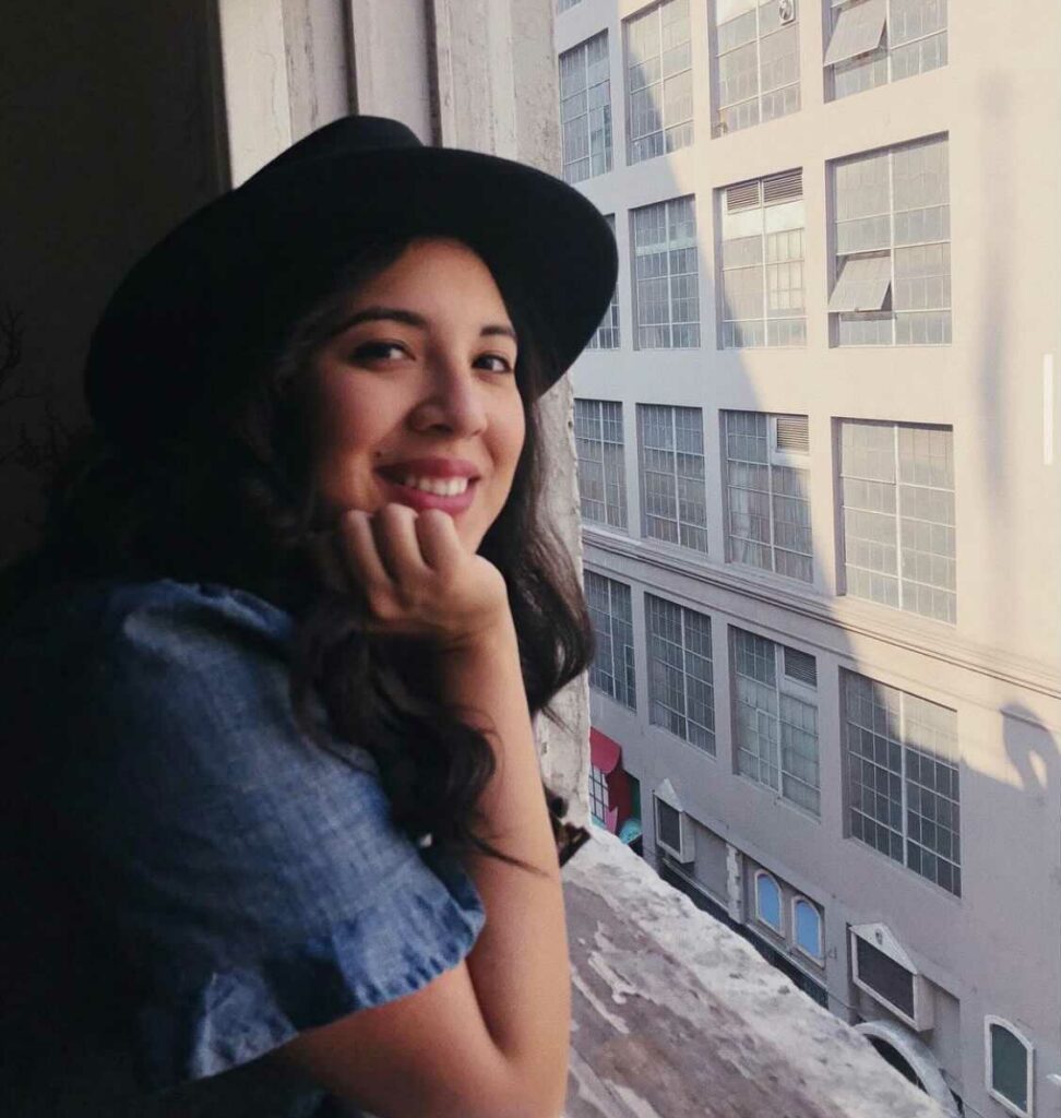 Berenice Santos smiling in a cowboy hat, leaning against a window